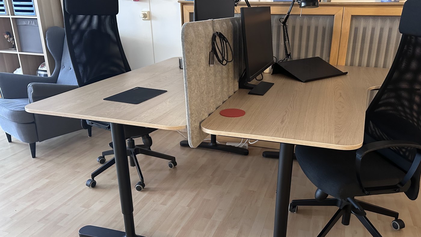 Blick in den Raum von der anderen Seite: zwei Schreibtische mit Filz-Sichtschutz, dahinter ein Sideboard, links ein Sessel.